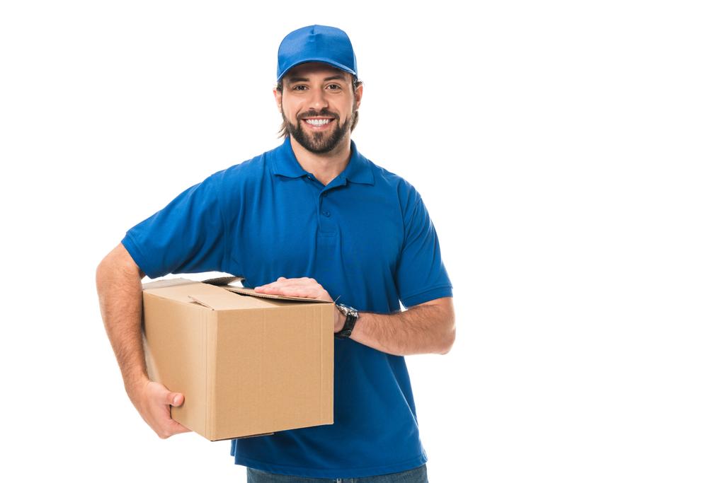 красивий молодий чоловік доставки тримає картонну коробку і посміхається на камеру ізольовано на білому
 - Фото, зображення
