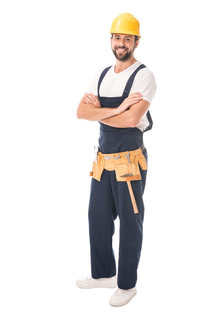 volle lengte weergave voor knappe gelukkig workman in harde hoed en gereedschap riem staande met gekruiste armen en lachend op camera geïsoleerd op wit - Foto, afbeelding