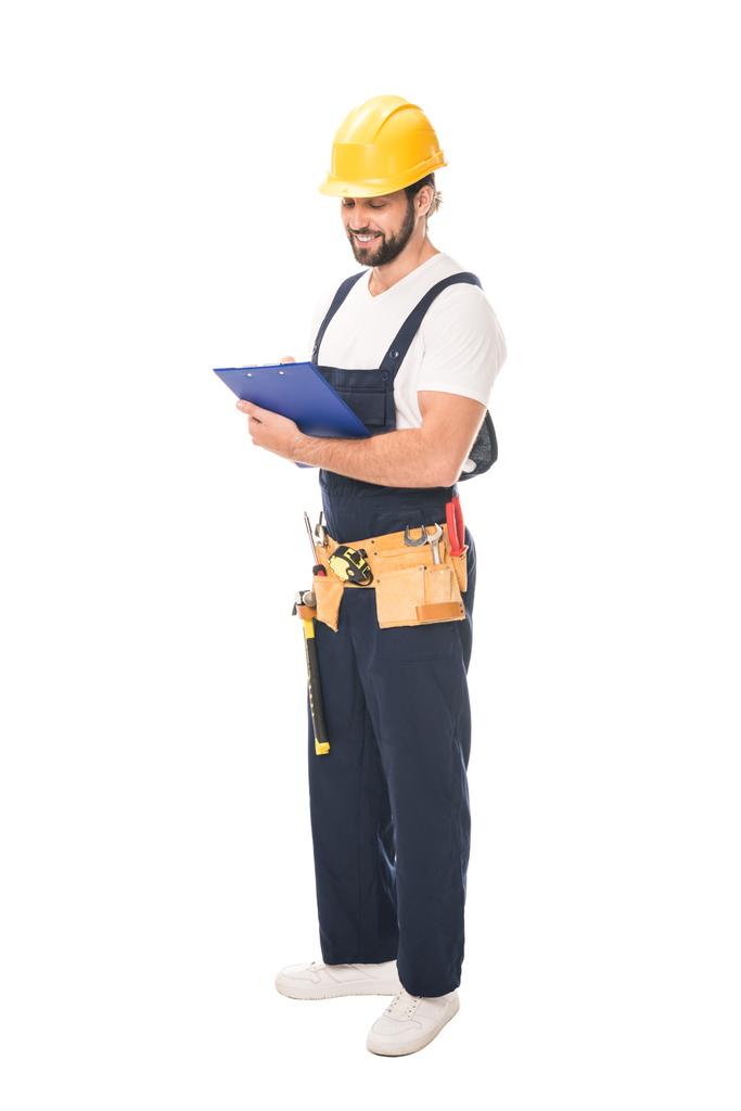 volle lengte weergave van glimlachen workman in gereedschap gordel schrijven op Klembord geïsoleerd op wit - Foto, afbeelding