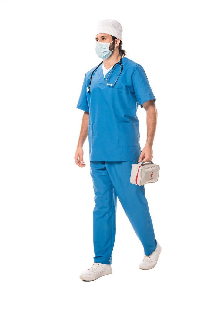 vista completa del médico en máscara médica sosteniendo botiquín de primeros auxilios y caminando aislado en blanco
 - Foto, Imagen