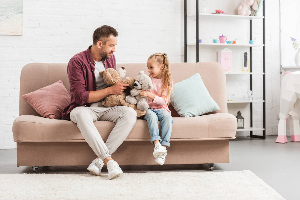 pai e filha brincando com ursos de pelúcia, sentados no sofá e olhando um para o outro
 - Foto, Imagem