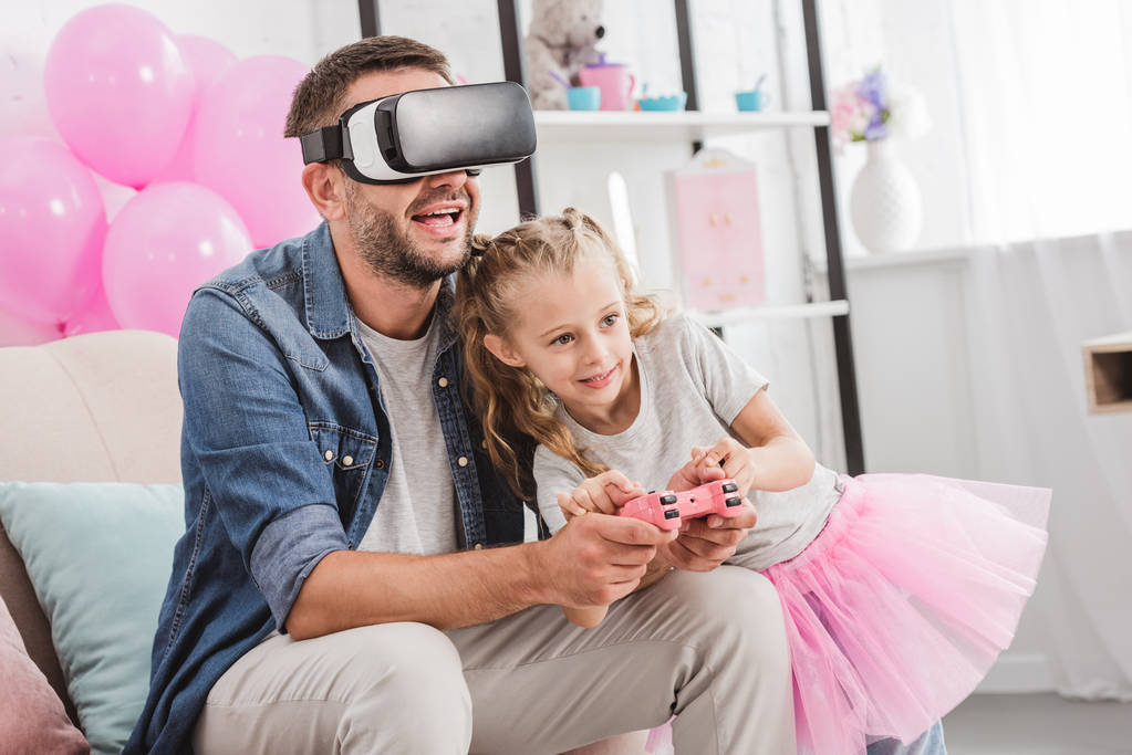 cheerful dad and daughter having fun and playing with joystick and using virtual reality headset on sofa - Photo, Image