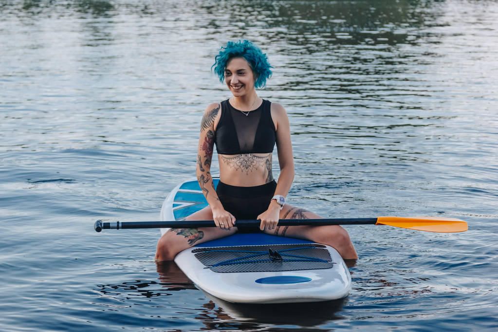 smiling girl with blue hair sitting on paddle board on river - Photo, Image