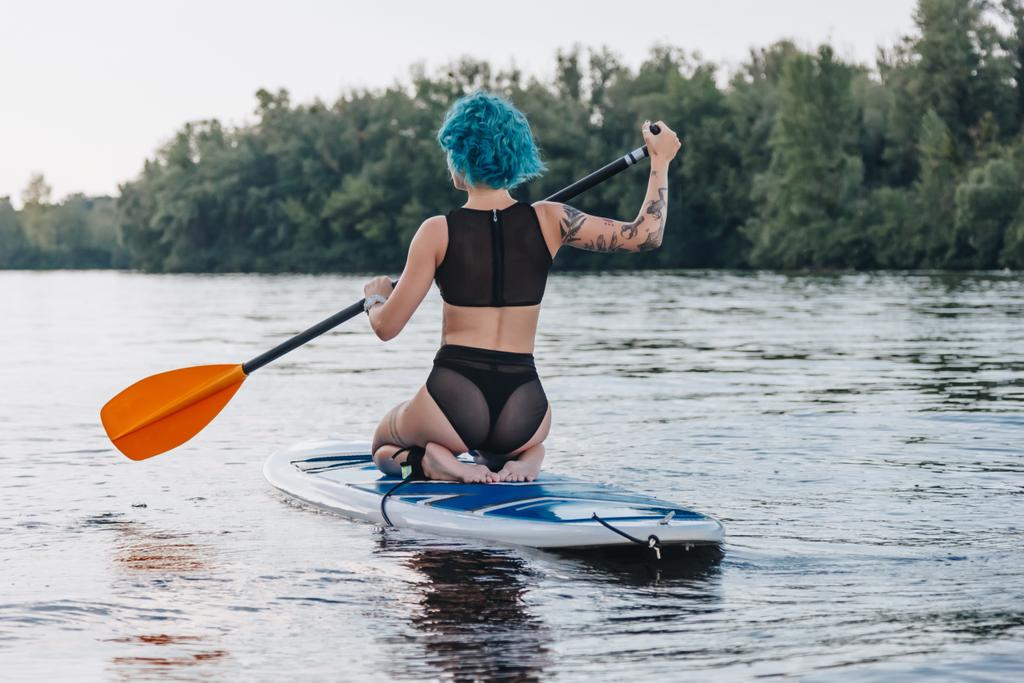 Вид сзади девушки с синими волосами, сидящей на доске на реке
 - Фото, изображение