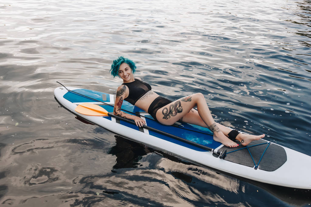 привлекательная татуированная девушка с голубыми волосами, лежащая на гребной доске на реке
 - Фото, изображение