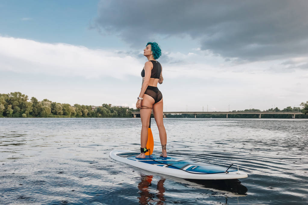 mooie sportieve jonge vrouw met blauwe haar standup paddleboarding op rivier  - Foto, afbeelding