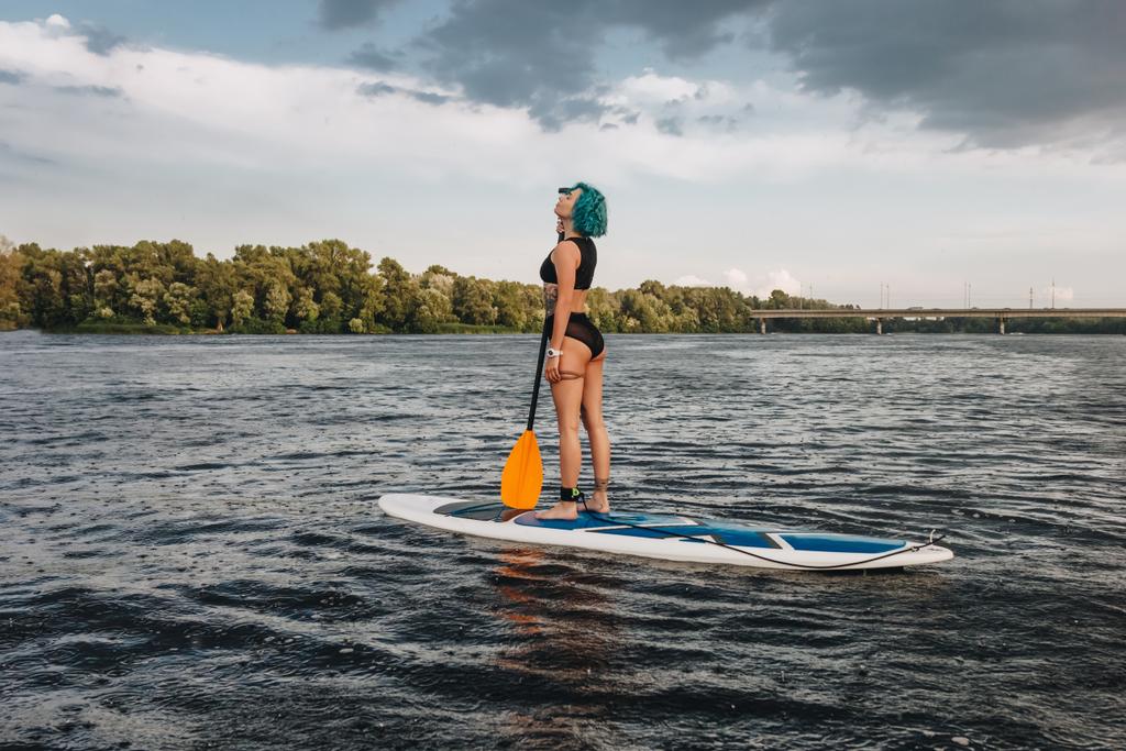 happy tattooed girl in bikini with blue hair paddle boarding on river - Photo, Image