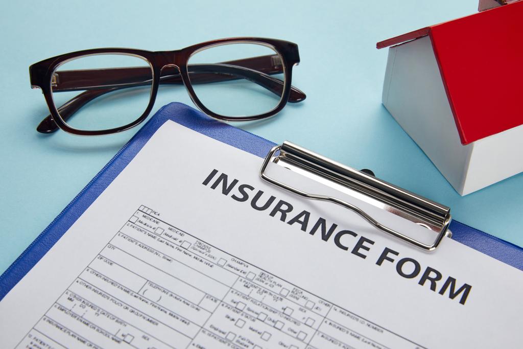 vergrote weergave van verzekeringsformulier op Klembord, brillen en huis model op blauw - Foto, afbeelding