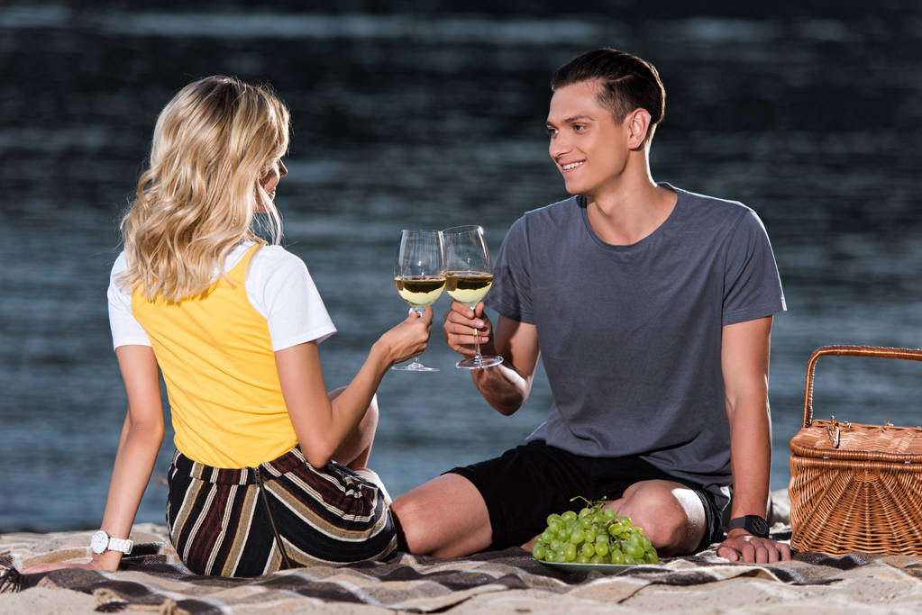 Seitenansicht eines jungen Paares, das bei einem Picknick am Flussstrand am Abend mit Weingläsern klingelt - Foto, Bild