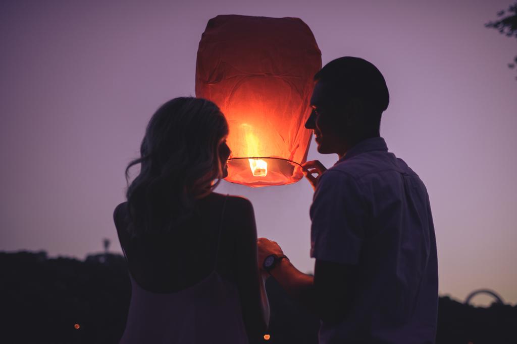 silhouettes of couple launching sky lantern on river beach in evening and looking at each other - Photo, Image