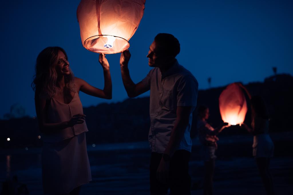 happy young couple launching sky lantern on river beach in evening - Photo, Image