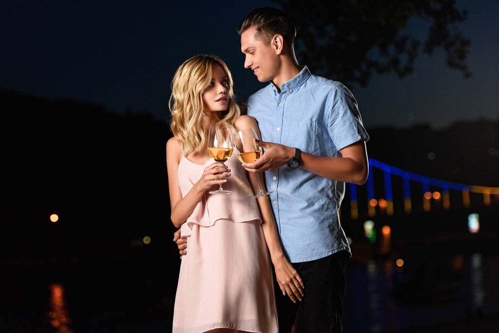 joven pareja tintineo con copas de vino y mirando unos a otros en la playa del río por la noche
 - Foto, imagen