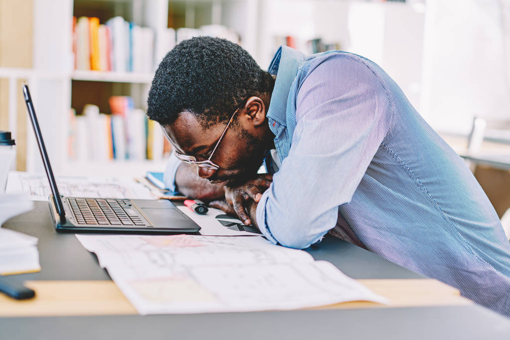 不幸なアフリカ系アメリカ人の男性の学生プロジェクトのデスクトップで座っていると、問題解決に集中して、オフィスでラップトップ コンピューターを介してオンラインで作業中に疲れている感じ暗い皮を剥がれた仕事中毒 - 写真・画像