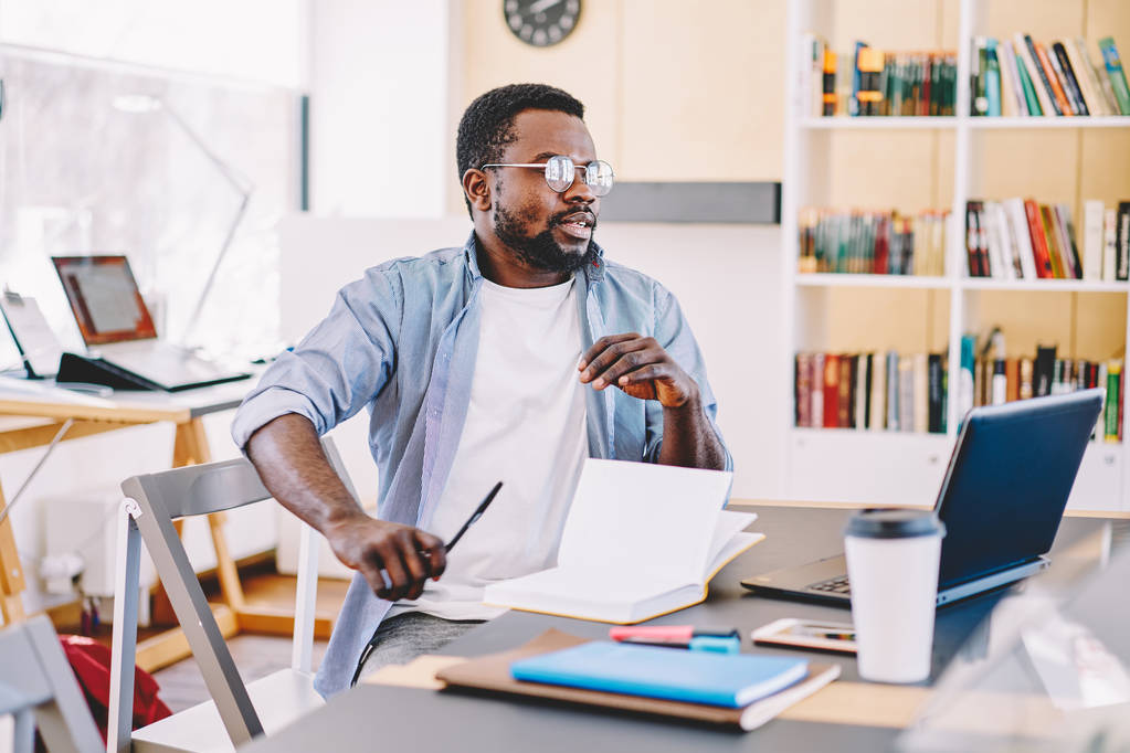 Ciddi koyu tenli erkek freelancer dizüstü bilgisayar, teknoloji ve wifi kitaplığında kullanarak ev ödevi görevi yapıyor akıllı Afro-Amerikan öğrenci ile masada oturan yayın için fikir üzerinde durulması - Fotoğraf, Görsel