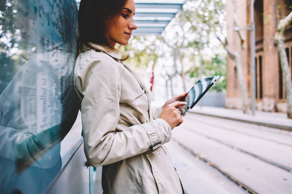 Вид сбоку стильной молодой женщины в трикотажном пальто, которая читает текст на цифровом планшете, подключенном к интернету 4G, стоя на трамвайной остановке в городской местности.
 - Фото, изображение