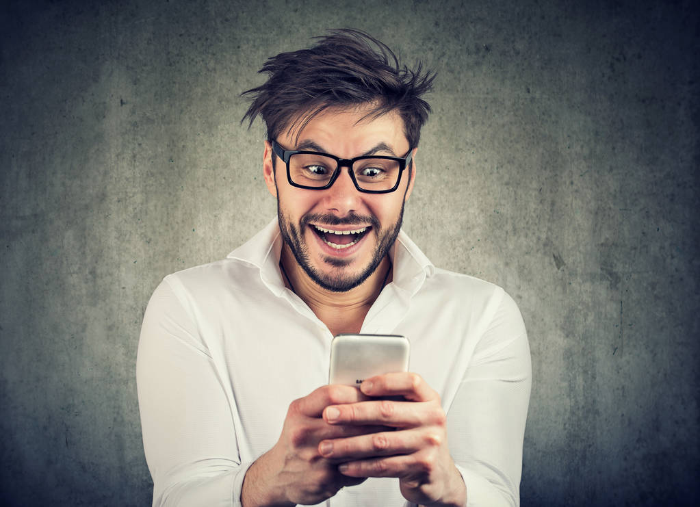 Вражаючий і серйозно виглядає молодий чоловік в окулярах і сорочках дивитися новини на смартфоні, що стоїть на сірому фоні
 - Фото, зображення