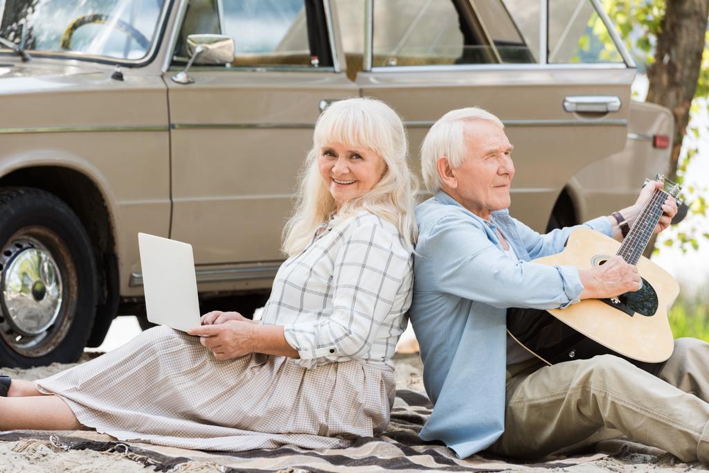 mujer mayor que utiliza el ordenador portátil mientras que el hombre tocando la guitarra contra coche vintage
  - Foto, Imagen
