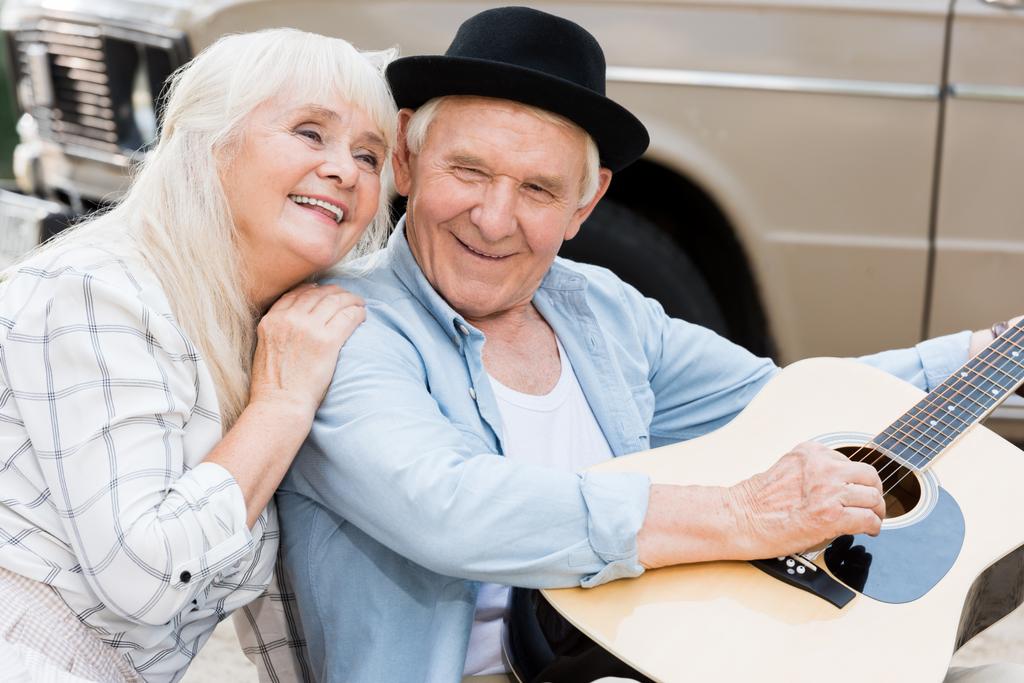 beautiful senior woman hugging man playing guitar against beige vintage car - Photo, Image