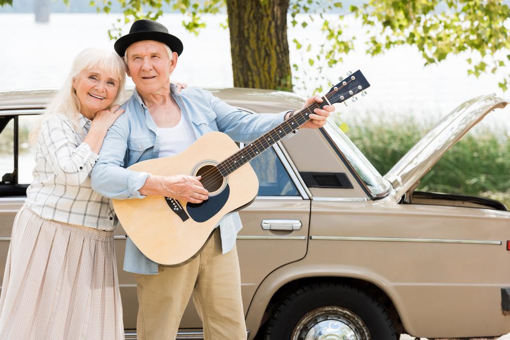 attrayant senior femme embrassant homme jouer de la guitare contre beige voiture vintage
 - Photo, image