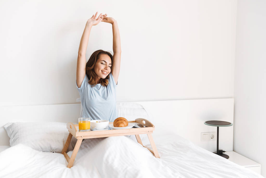 Uśmiechający się młoda kobieta, rozciąganie jej ręce, jedząc śniadanie na tacy w łóżku - Zdjęcie, obraz