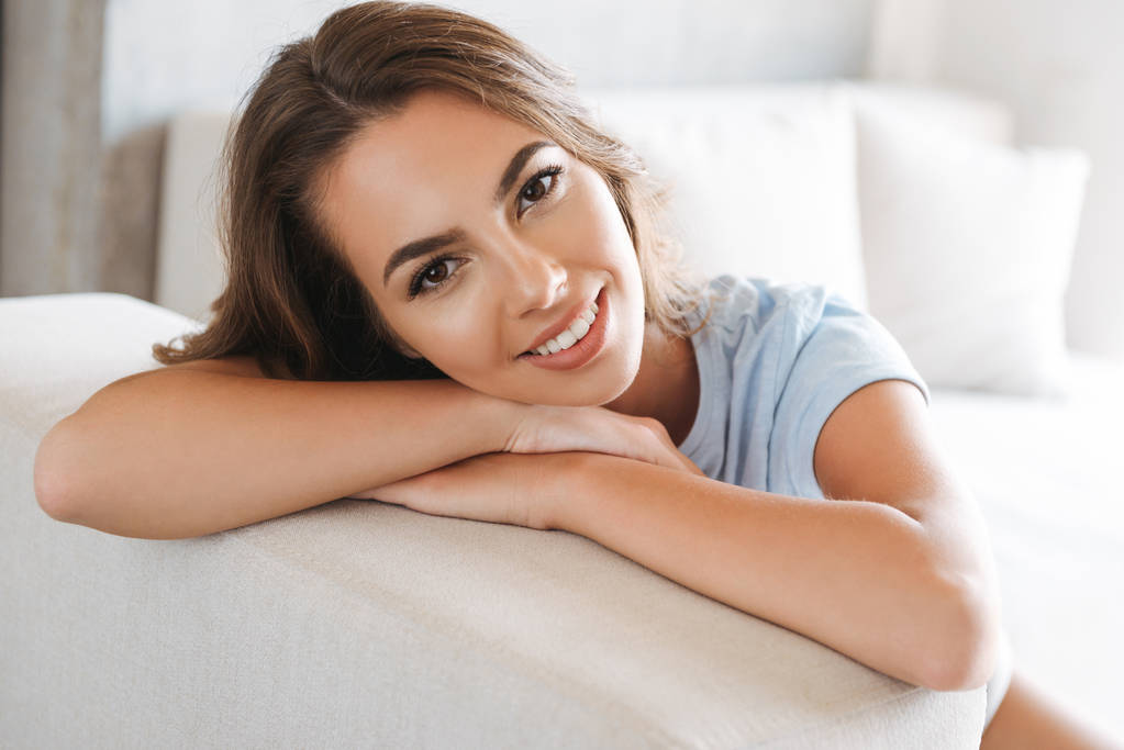 Lähikuva hymyilevä nuori nainen rentoutuu sohvalla kotona ja katselee kameraa
 - Valokuva, kuva