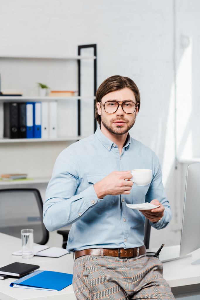 красивый молодой бизнесмен с чашкой кофе, смотрящий в камеру в современном офисе
 - Фото, изображение