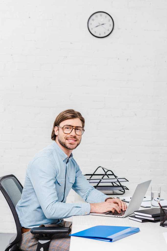 sorridente jovem empresário trabalhando com laptop no escritório moderno e olhando para a câmera
 - Foto, Imagem