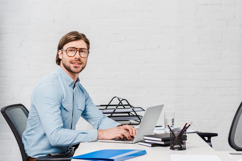 jovem empresário feliz trabalhando com laptop no escritório moderno e olhando para a câmera
 - Foto, Imagem