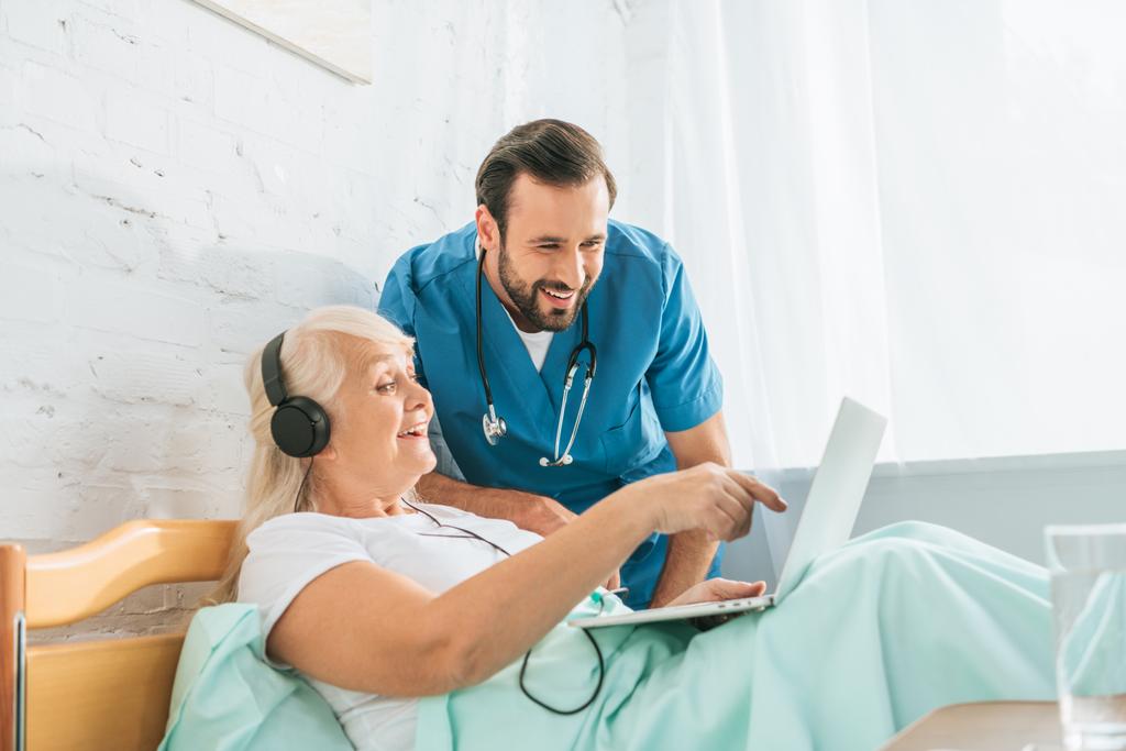усміхнена чоловіча медсестра дивиться на щасливу старшу жінку в навушниках, використовуючи ноутбук у лікарняному ліжку
    - Фото, зображення