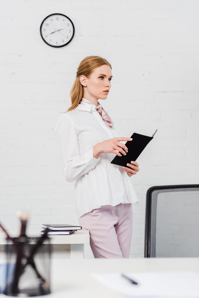 confident young businesswoman with notebook standing at modern office and looking away - Photo, Image
