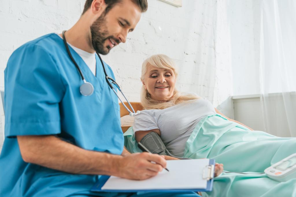 усміхнена чоловіча медсестра пише в буфері, вимірюючи кров'яний тиск на старшу жінку, що лежить у ліжку
 - Фото, зображення