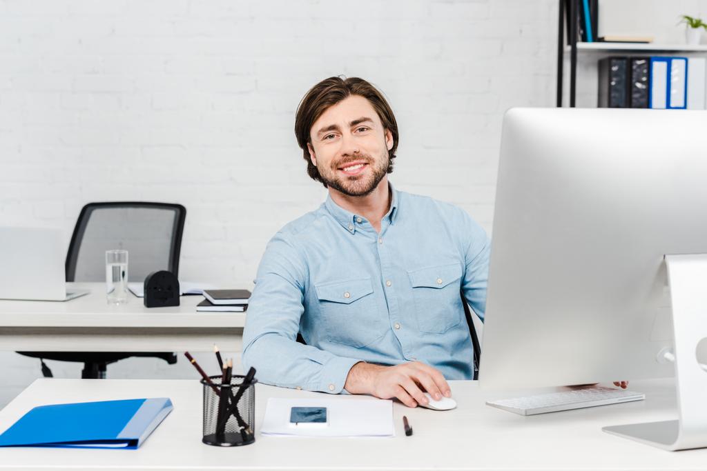 mosolygó fiatal üzletember keresi a kamera munka közben a számítógép modern irodában - Fotó, kép
