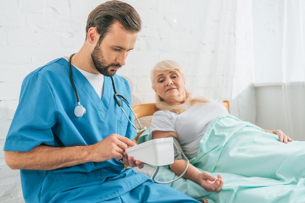 молода чоловіча медсестра зі стетоскопом, що тримає монітор артеріального тиску при вимірюванні кров'яного тиску для старшої жінки, що лежить у ліжку
 - Фото, зображення