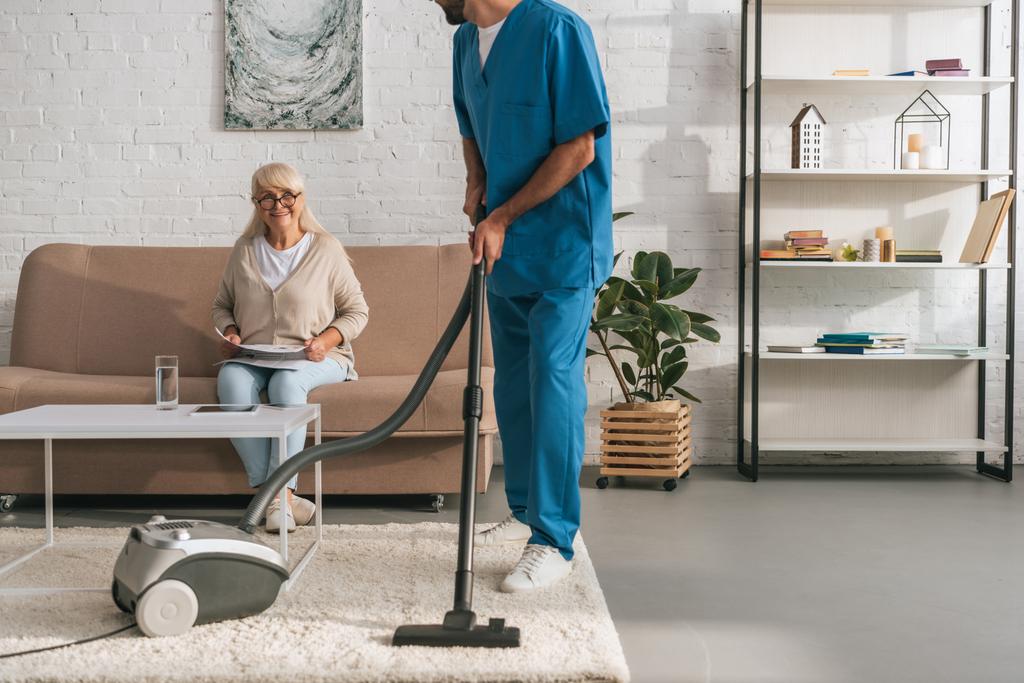 Schnappschuss von Sozialarbeiterin, die Teppich mit Staubsauger reinigt, während Seniorin Zeitung liest - Foto, Bild