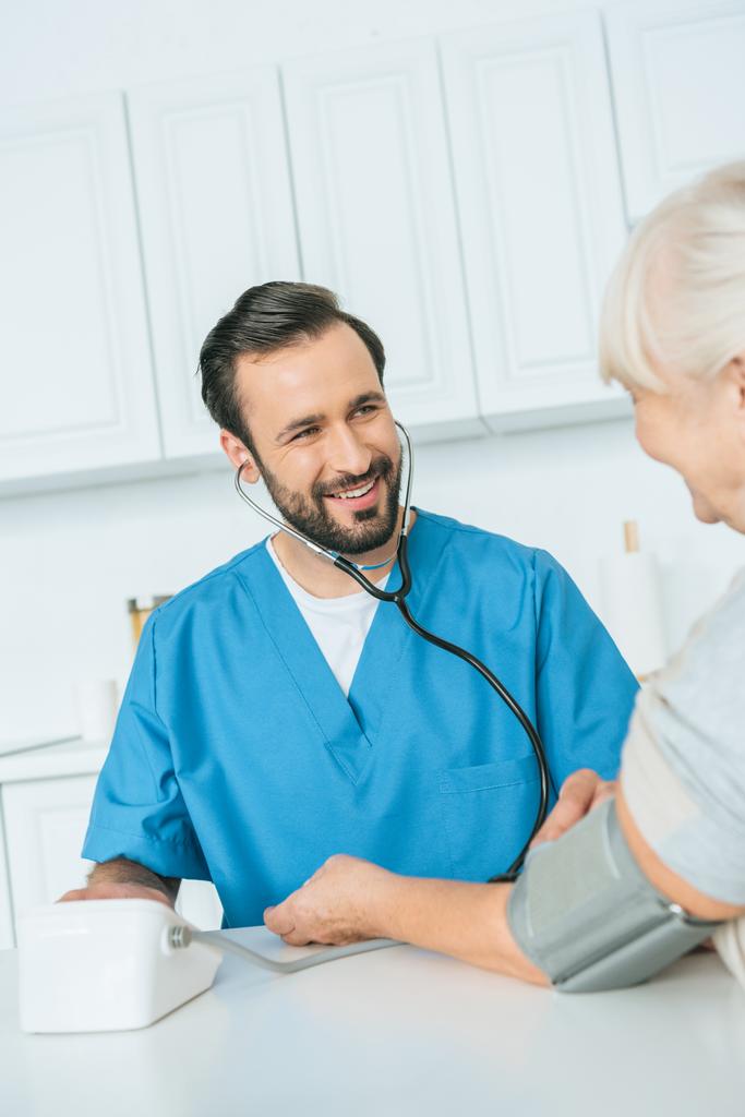 年配の女性に血圧測定のオスの看護婦の笑顔のショットをトリミング - 写真・画像
