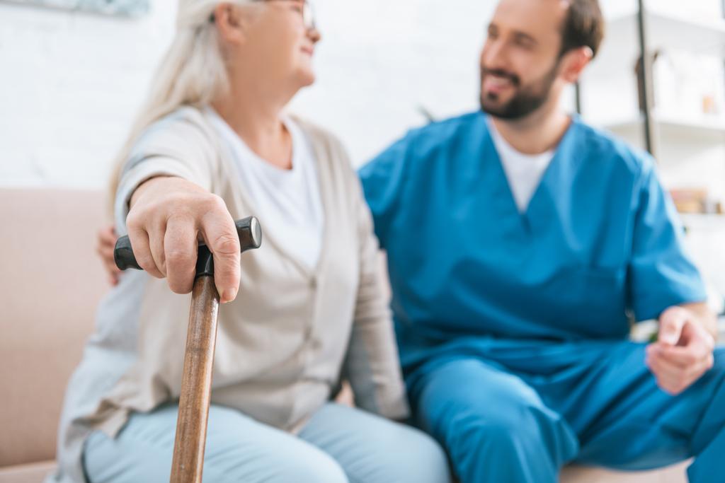 close-up vista da mulher idosa segurando bengala andando e olhando para enfermeira masculina sorridente
  - Foto, Imagem