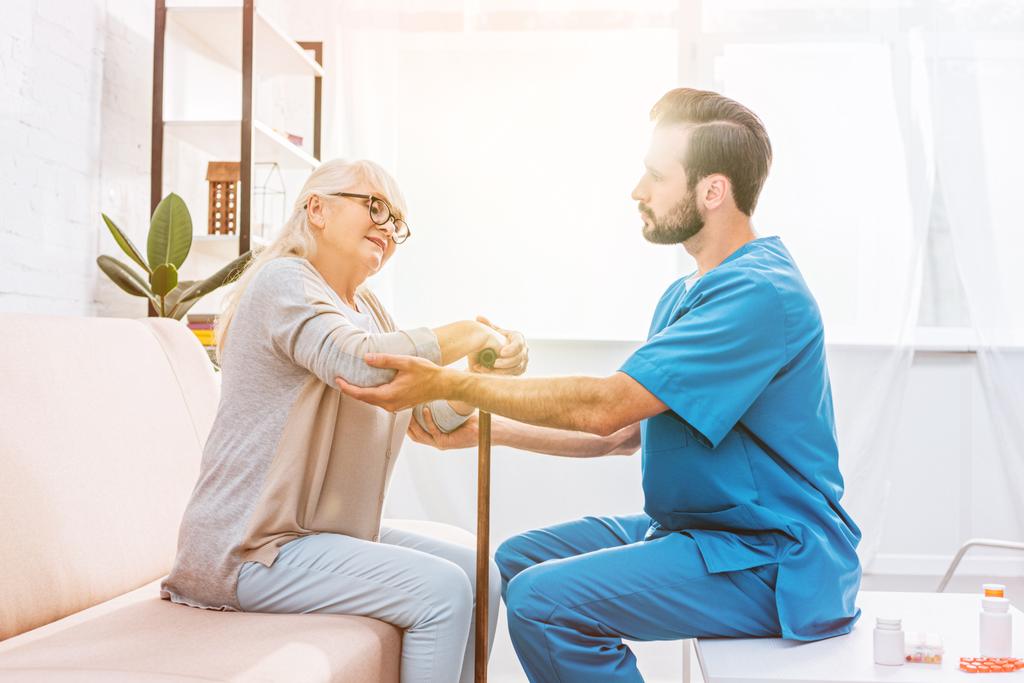 vista lateral de la enfermera masculina que apoya a la mujer mayor con bastón
 - Foto, imagen