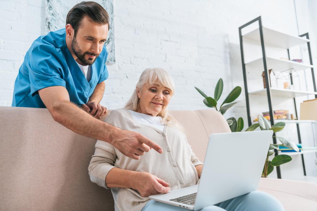 młody mężczyzna pracownik socjalny, wskazując palcem na laptop biały starszy kobieta siedzi na kanapie - Zdjęcie, obraz