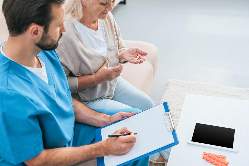visão de alto ângulo da enfermeira masculina escrevendo na área de transferência e mulher idosa tomando pílulas
 - Foto, Imagem