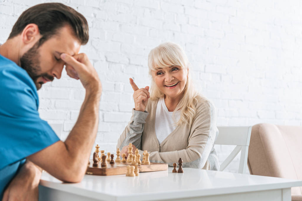 Lächelnde Seniorin zeigt mit dem Finger und blickt aufgebrachten Mann nach Schachspiel an - Foto, Bild