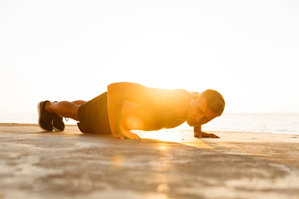 Concentrated sportsman with earphones doing push-ups at the beach in sunlight - Photo, Image