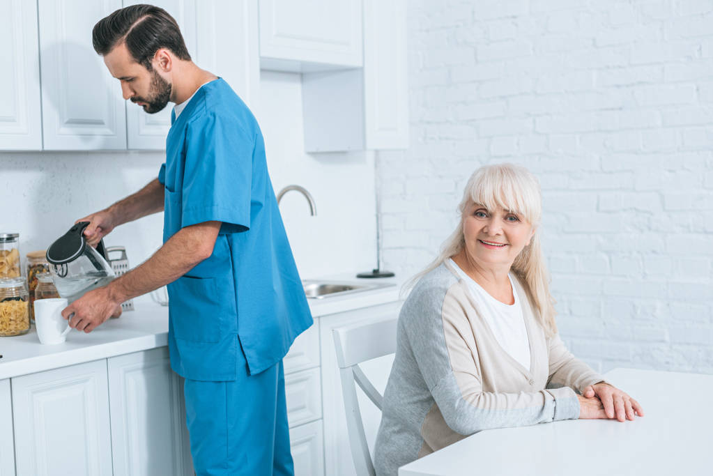 Glückliche Seniorin sitzt lächelnd in die Kamera, während Betreuerin Heißgetränk in Küche einschenkt - Foto, Bild