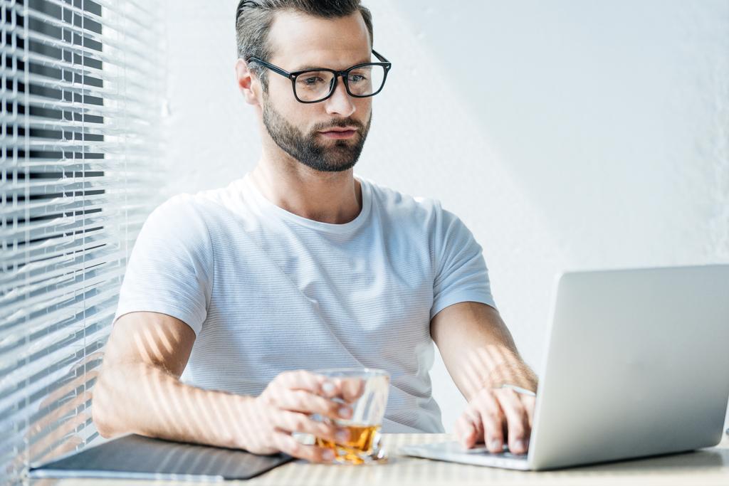 Брюнет мужчина держит стакан виски и работает с ноутбуком
 - Фото, изображение