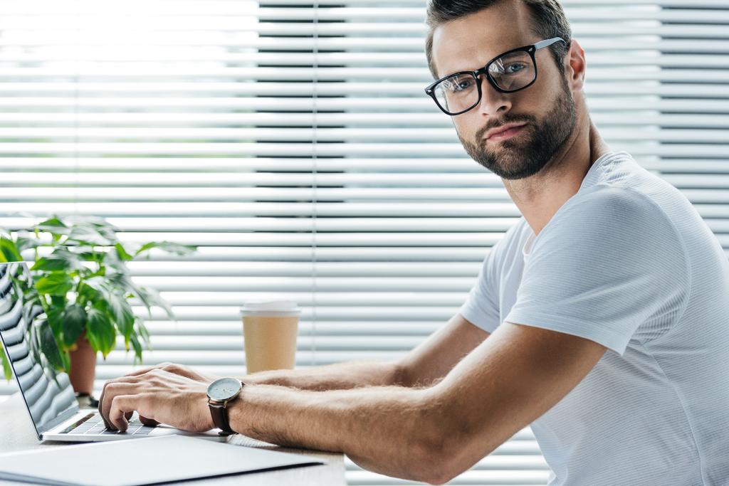 бородатий чоловік використовує ноутбук, сидячи на робочому місці з одноразовою чашкою кави
 - Фото, зображення