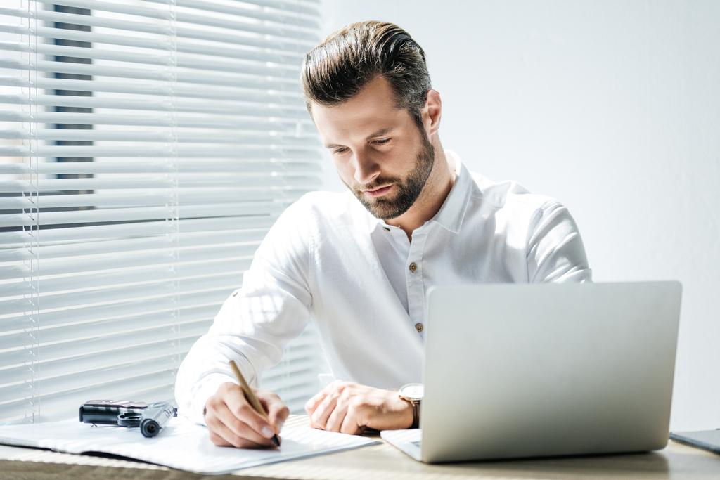 concentrated businessman writing in documents while sitting at workplace with laptop and gun - Photo, Image