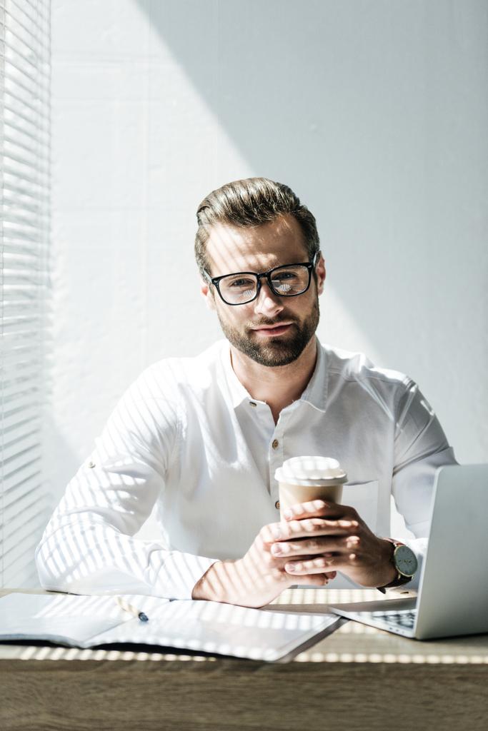 pohledný podnikatel sedí na pracovišti s notebookem, papírování a šálek kávy  - Fotografie, Obrázek
