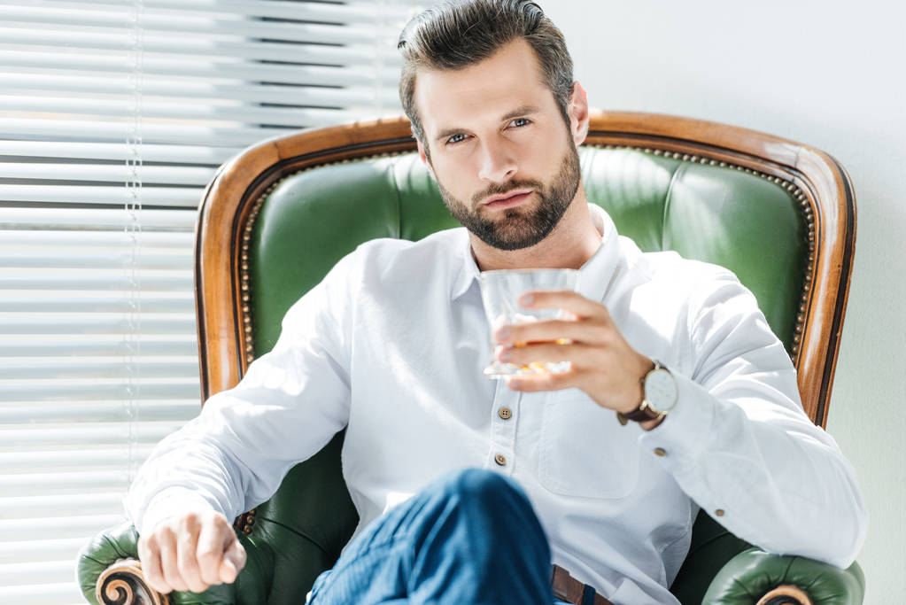 guapo elegante hombre sosteniendo vaso de whisky y sentado en un sillón verde
 - Foto, Imagen