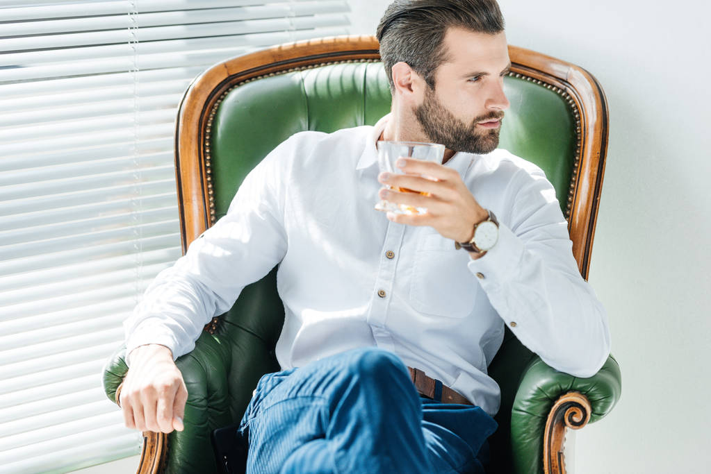 elegantní stylový podnikatel drží sklenici whisky a sedí v křesle - Fotografie, Obrázek