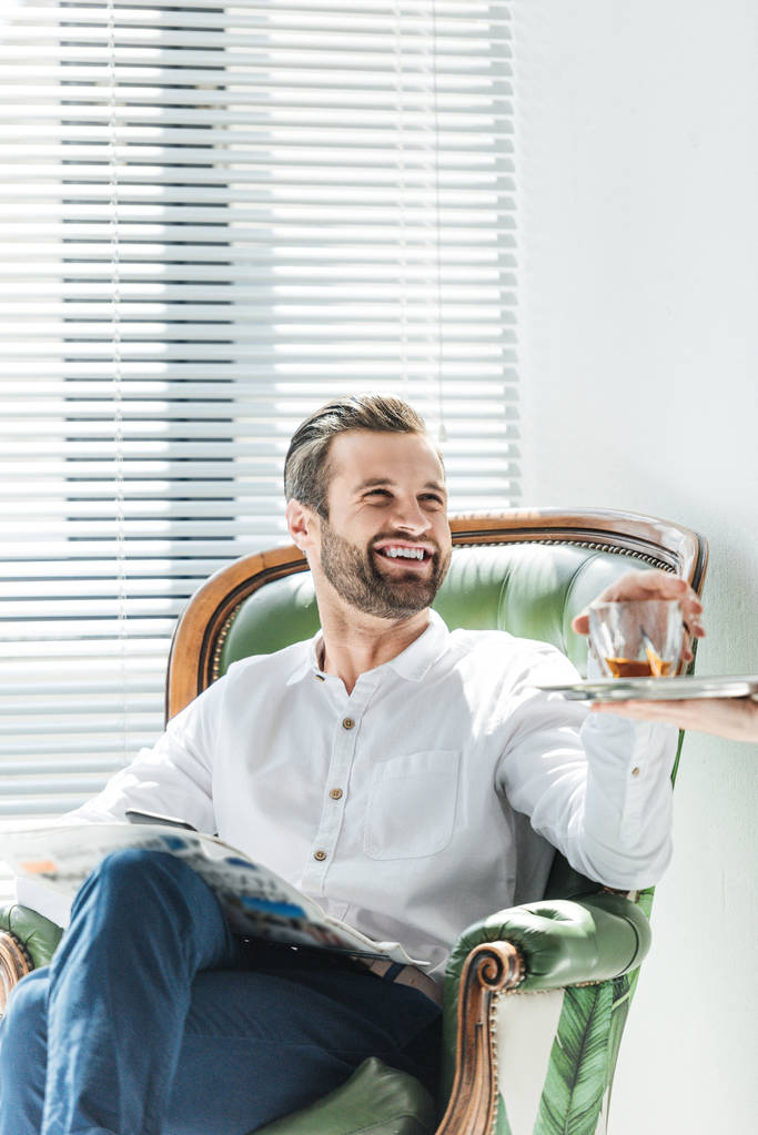 hombre sonriente guapo con periódico tomando vaso de whisky de la bandeja
 - Foto, imagen
