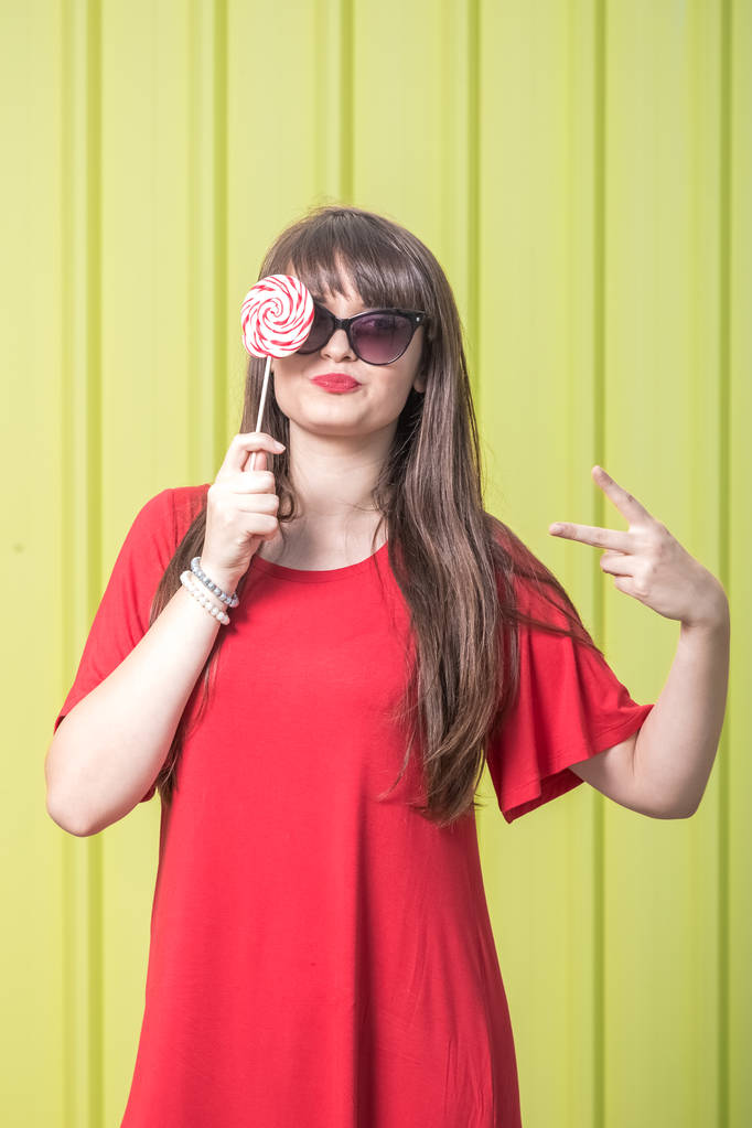 Cheerful girl with sunglasses in red dress covering her eye with lollipop candy against yellow background. - Photo, Image